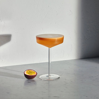 Modern clear glass coupe with a thin stem filled with an orange cocktail next to a halved passion fruit on marble.