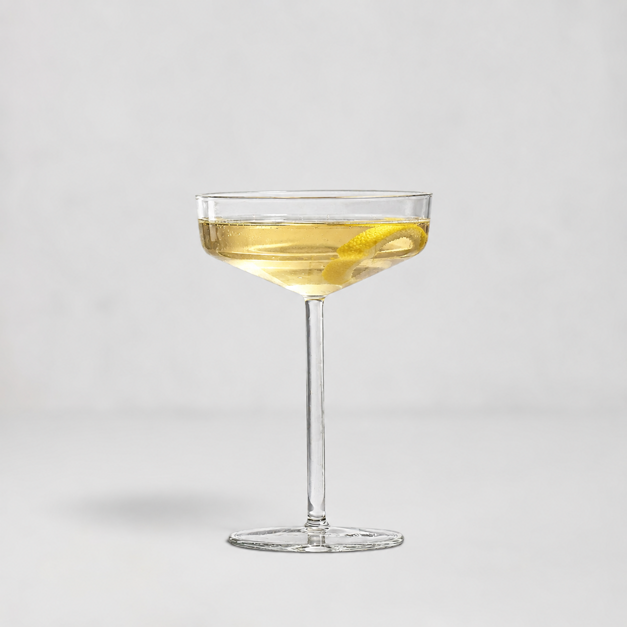 A clear coupe glass filled with a light golden drink and a lemon twist, centered on a clean light gray background.