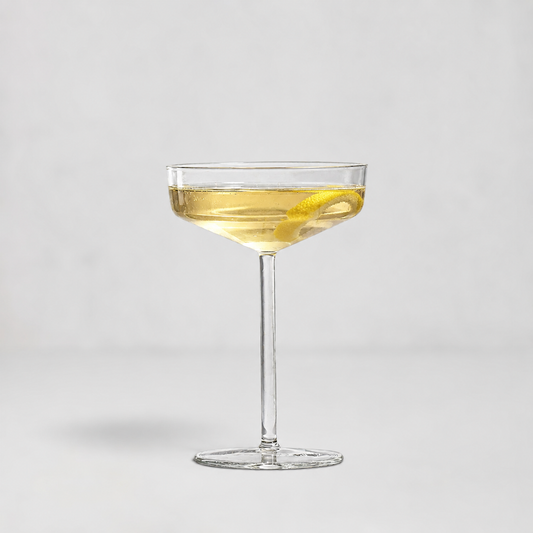 A clear coupe glass filled with a light golden drink and a lemon twist, centered on a clean light gray background.
