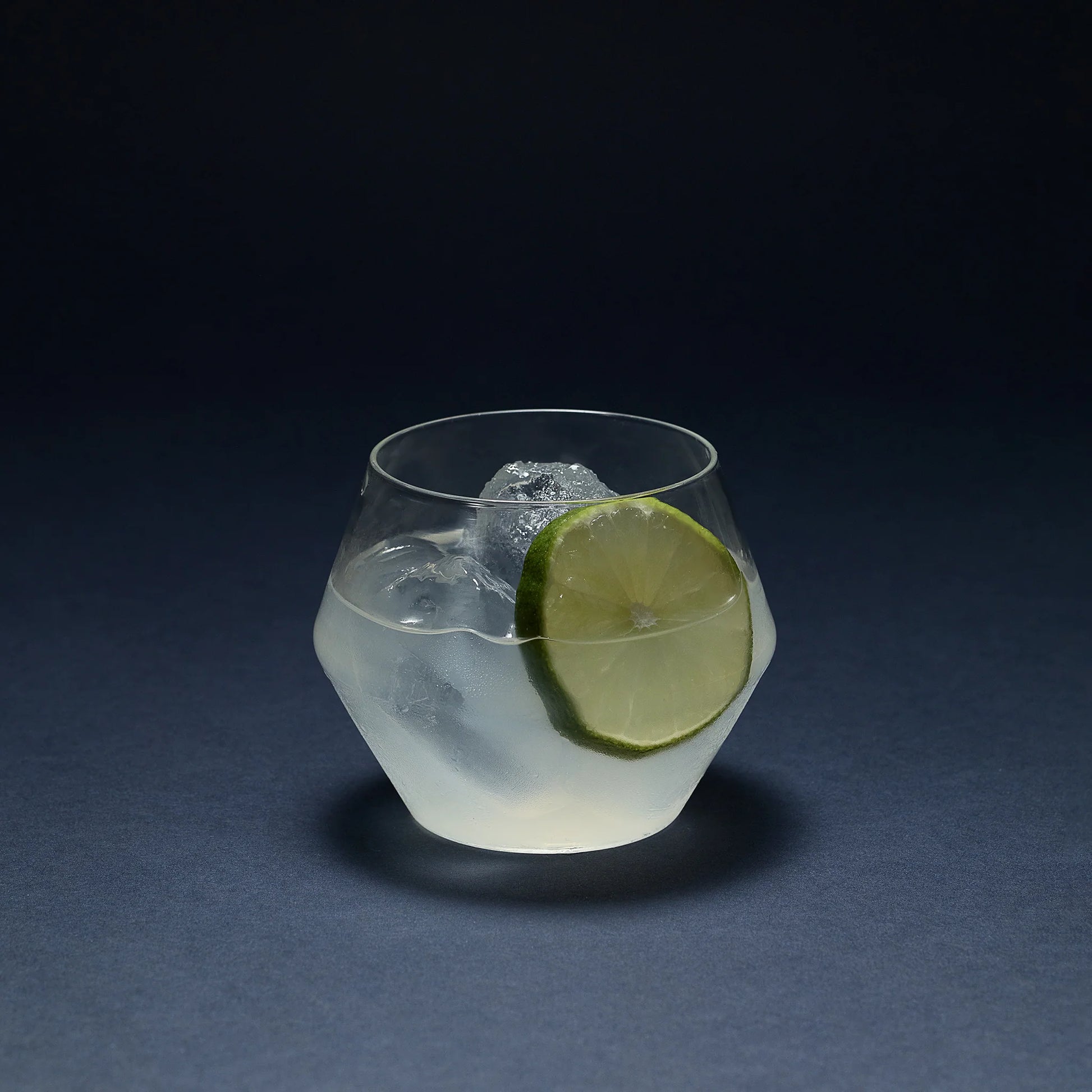 Clear glass Juniper double old fashioned tumbler with a geometric silhouette, containing a cocktail, ice, and lime slice.
