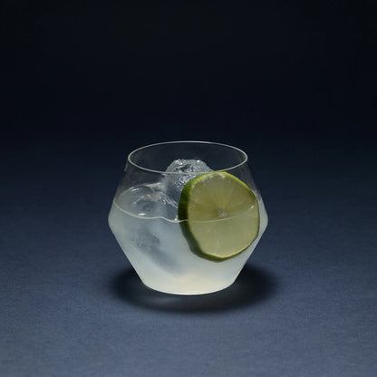 Clear glass Juniper double old fashioned tumbler with a geometric silhouette, containing a cocktail, ice, and lime slice.