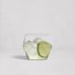 A clear lowball-style cocktail glass with a faceted hexagonal base, filled with ice and a lime slice, photographed on a clean light gray background.
