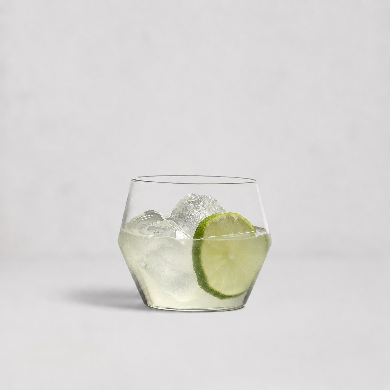 A clear lowball-style cocktail glass with a faceted hexagonal base, filled with ice and a lime slice, photographed on a clean light gray background.