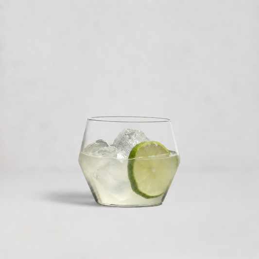 A clear lowball-style cocktail glass with a faceted hexagonal base, filled with ice and a lime slice, photographed on a clean light gray background.