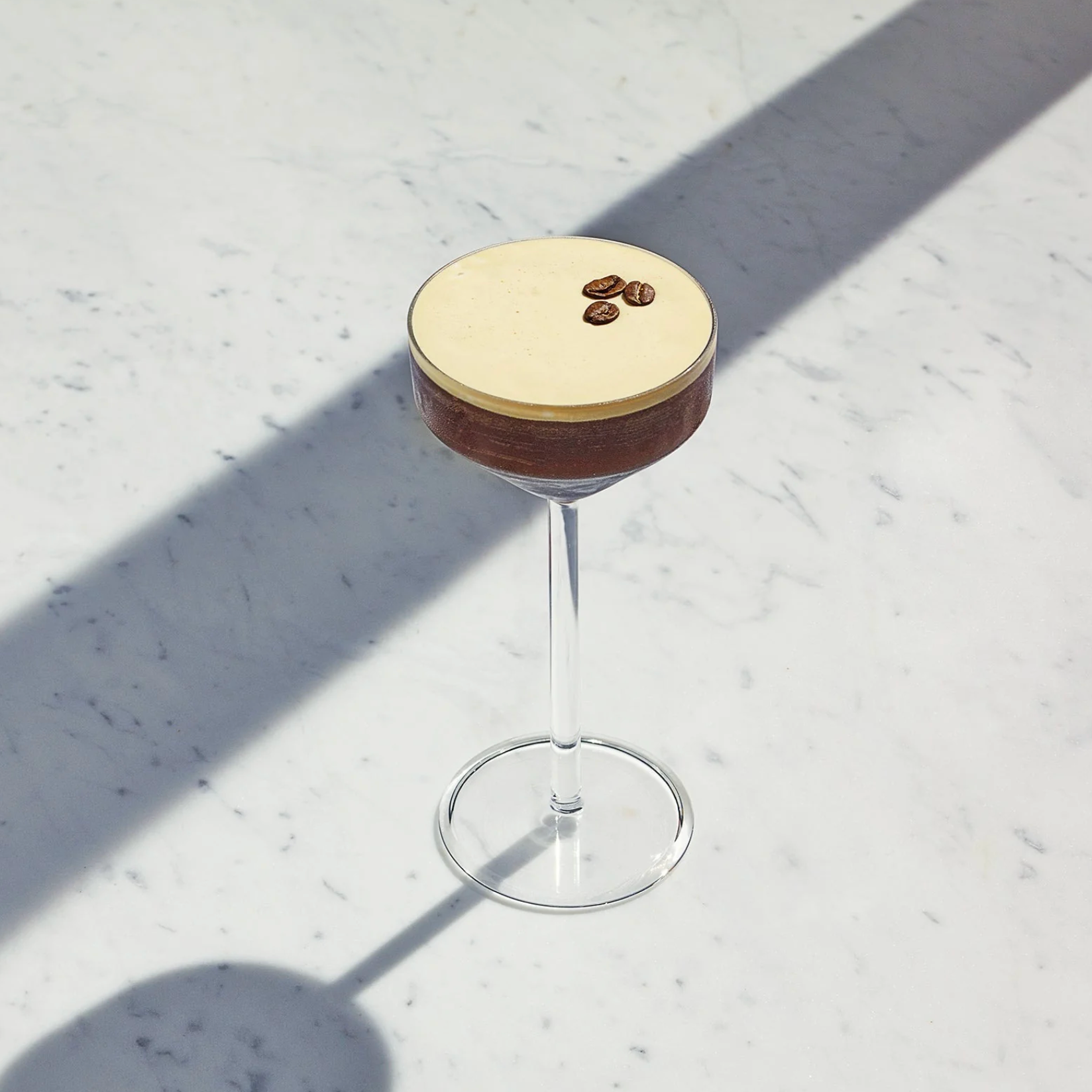 A layered espresso martini in a coupe glass, featuring a dark brown chocolate layer and a light beige cream layer on top, garnished with three coffee beans, set on a white marble surface with strong diagonal shadows.