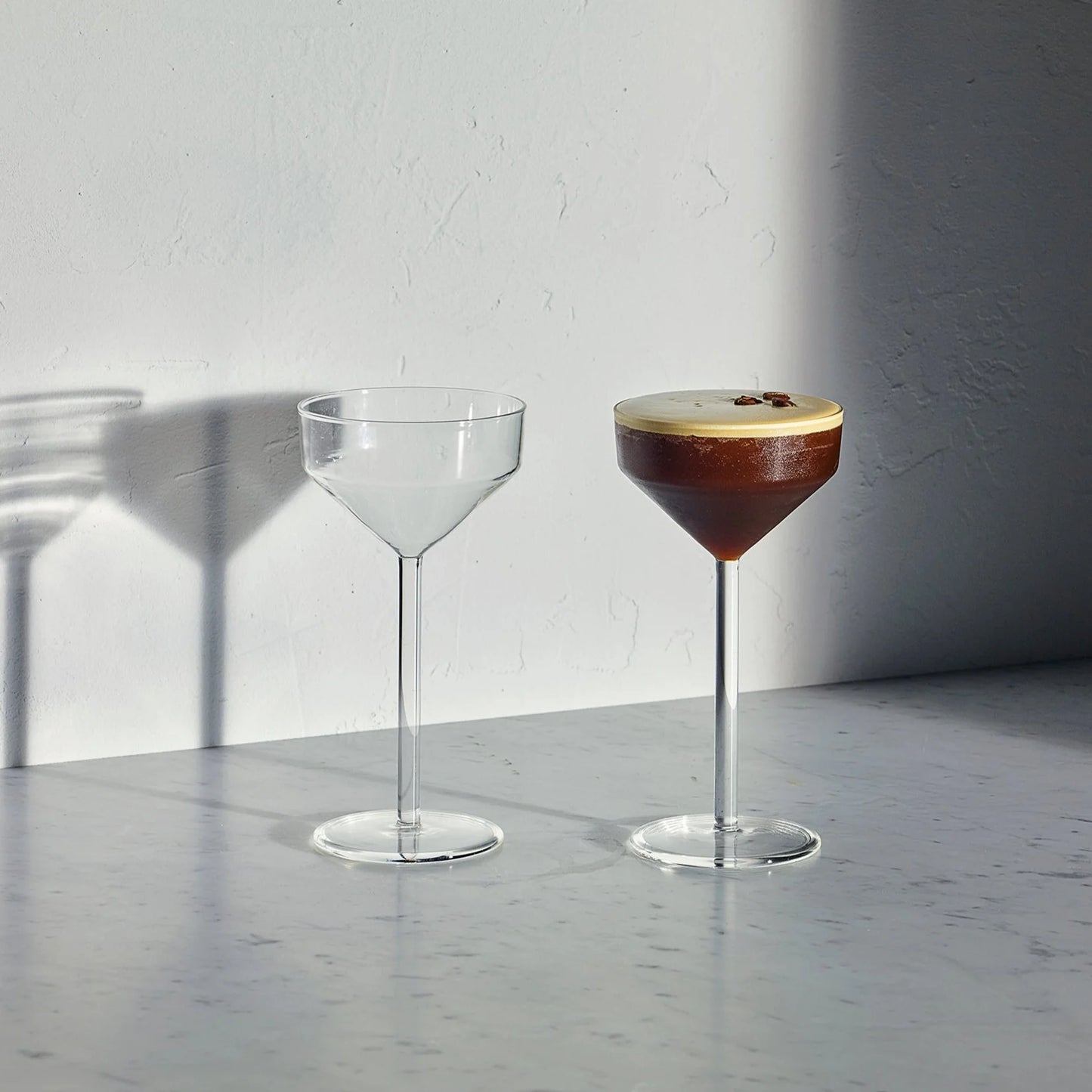 Two clear glass Juniper cocktail glasses with sculptural geometric bowls and slender stems on a marble surface.