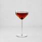 A clear stemmed coupe-style cocktail glass filled with an amber-red drink and a dark olive garnish, centered on a light gray studio background.
