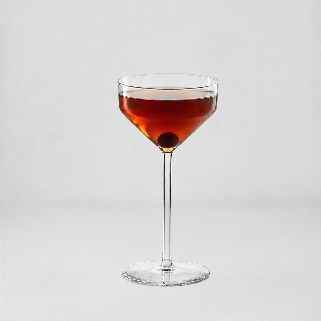 A clear stemmed coupe-style cocktail glass filled with an amber-red drink and a dark olive garnish, centered on a light gray studio background.