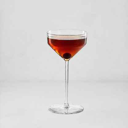 A clear stemmed coupe-style cocktail glass filled with an amber-red drink and a dark olive garnish, centered on a light gray studio background.