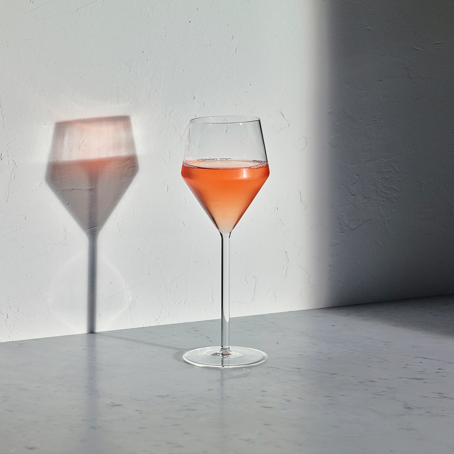 Juniper white wine glass with a geometric bowl and thin stem, filled with rose wine, casting a shadow on a white wall.