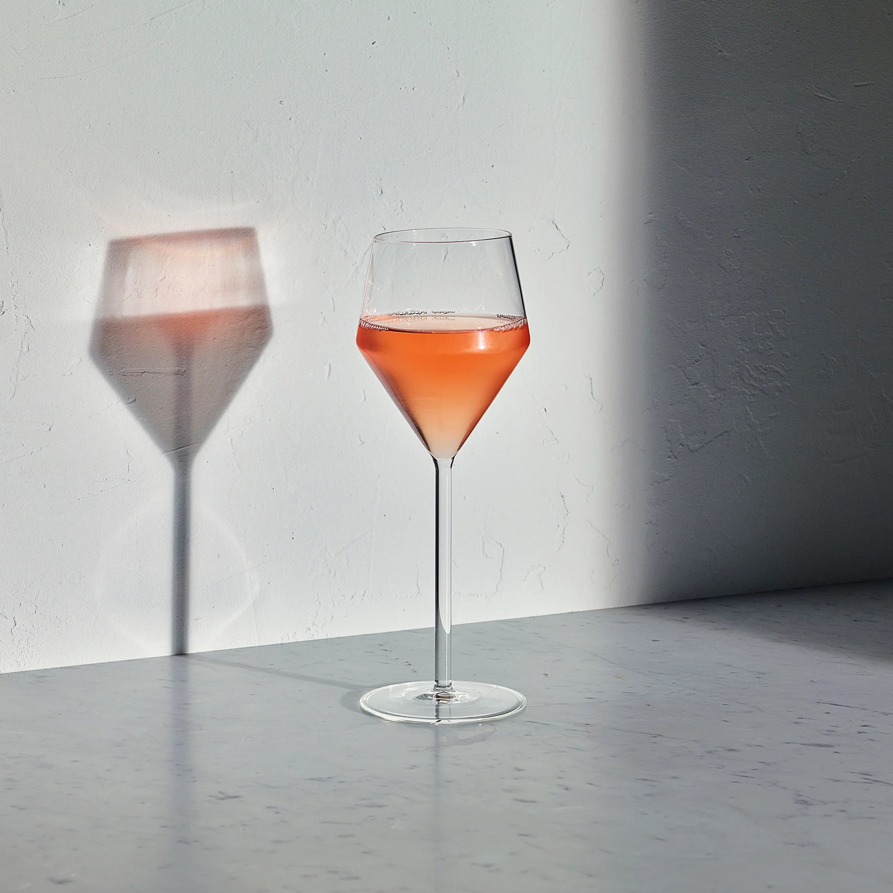 Juniper white wine glass with a geometric bowl and thin stem, filled with rose wine, casting a shadow on a white wall.