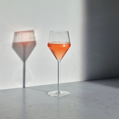 Juniper white wine glass with a geometric bowl and thin stem, filled with rose wine, casting a shadow on a white wall.