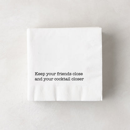 White 3-ply paper cocktail napkins with "Keep your friends close and your cocktail closer" in black text.