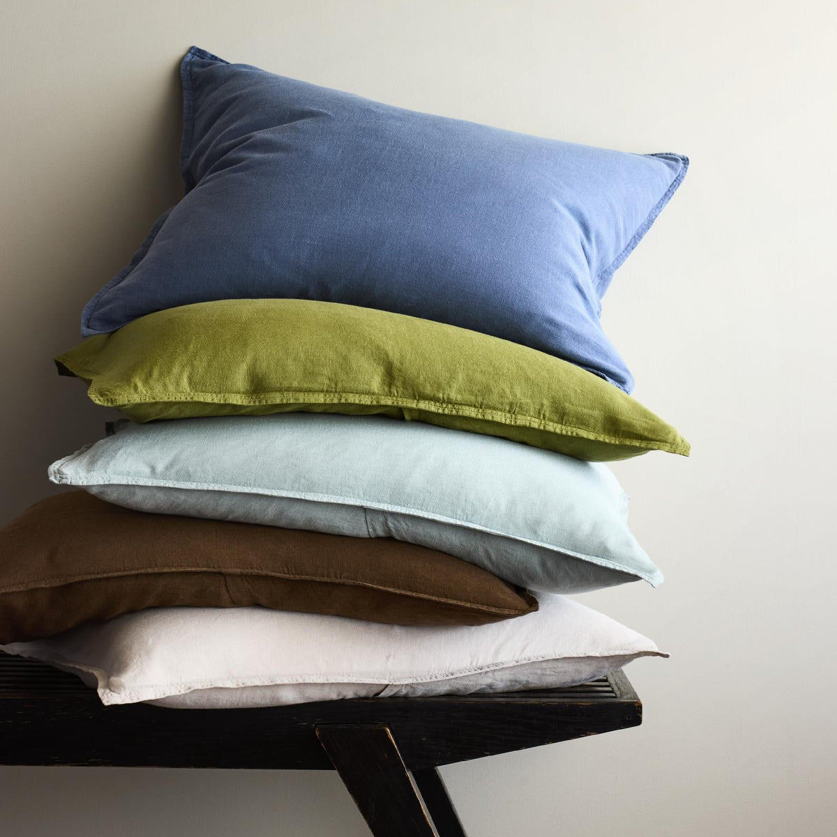 Stack of five washed linen pillow shams in blue, green, light blue, brown, and white on a black wooden bench.