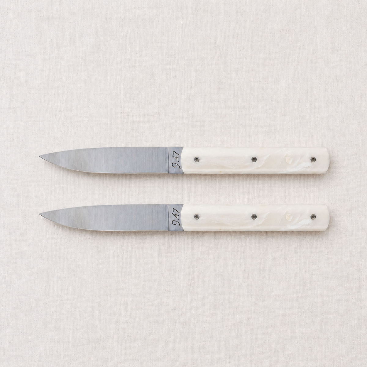 Set of two Perceval 9.47 steak knives with stainless steel blades and cream-colored Krion handles on a linen background.