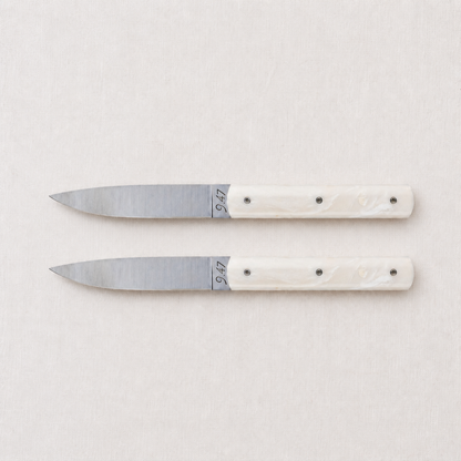 Set of two Perceval 9.47 steak knives with stainless steel blades and cream-colored Krion handles on a linen background.