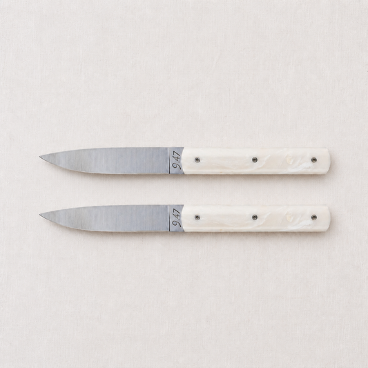 Set of two Perceval 9.47 steak knives with stainless steel blades and cream-colored Krion handles on a linen background.