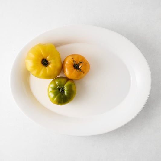 White oval ceramic Lagos serving platter with three heirloom tomatoes: yellow, orange, and green.