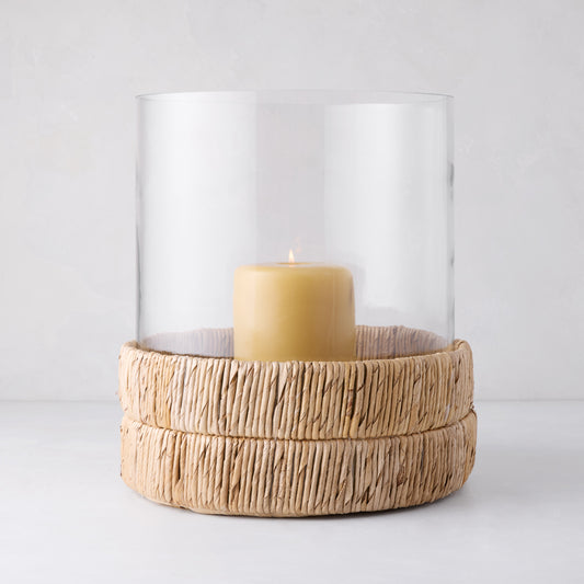 Large glass hurricane with a woven banana bark base and a lit amber candle.