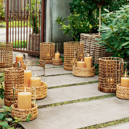 Large glass hurricane with woven hyacinth base, holding an amber candle, on a patio.