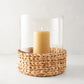 Large glass hurricane with woven water hyacinth base and lit amber candle.