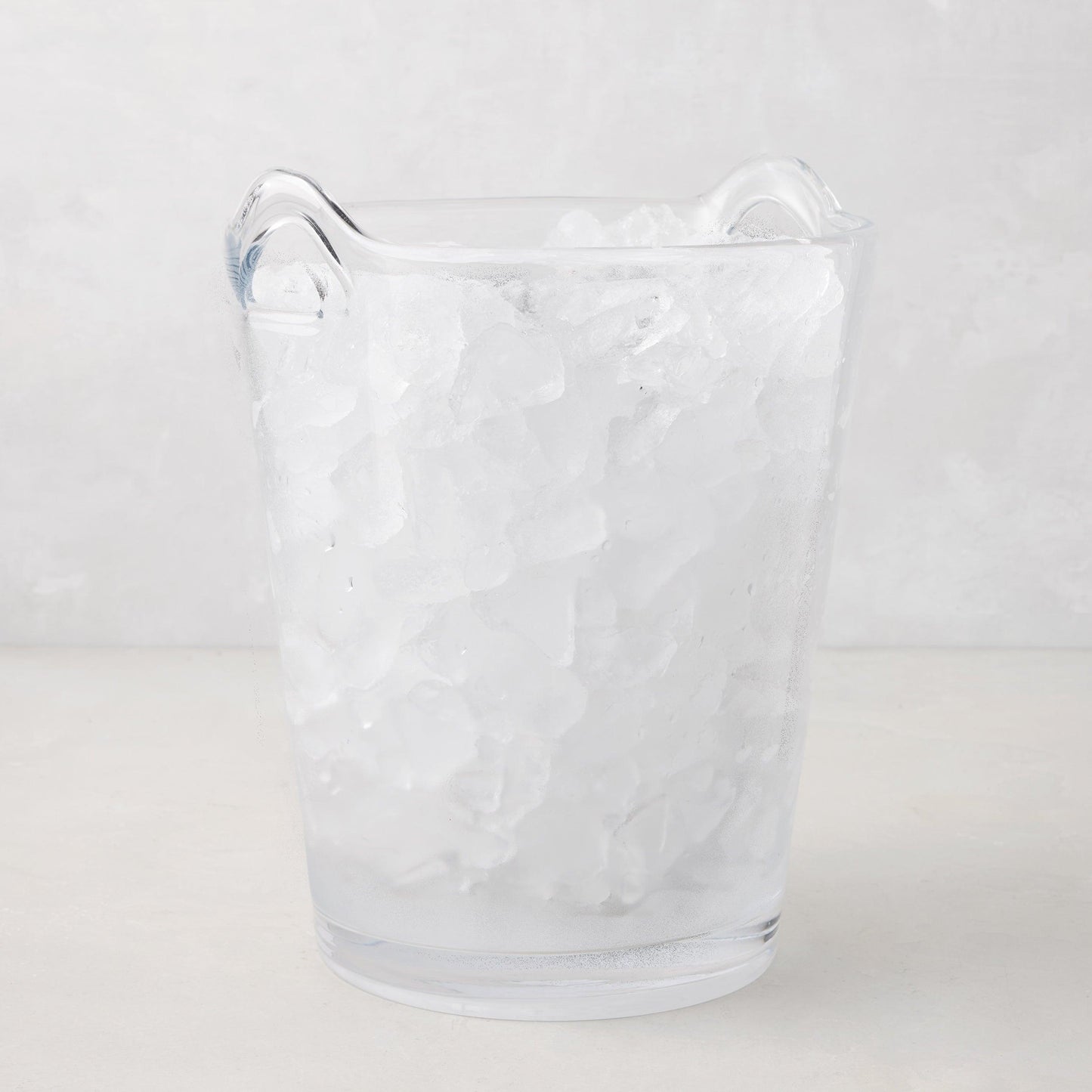 Large clear handblown glass ice bucket with two handles, filled with ice.