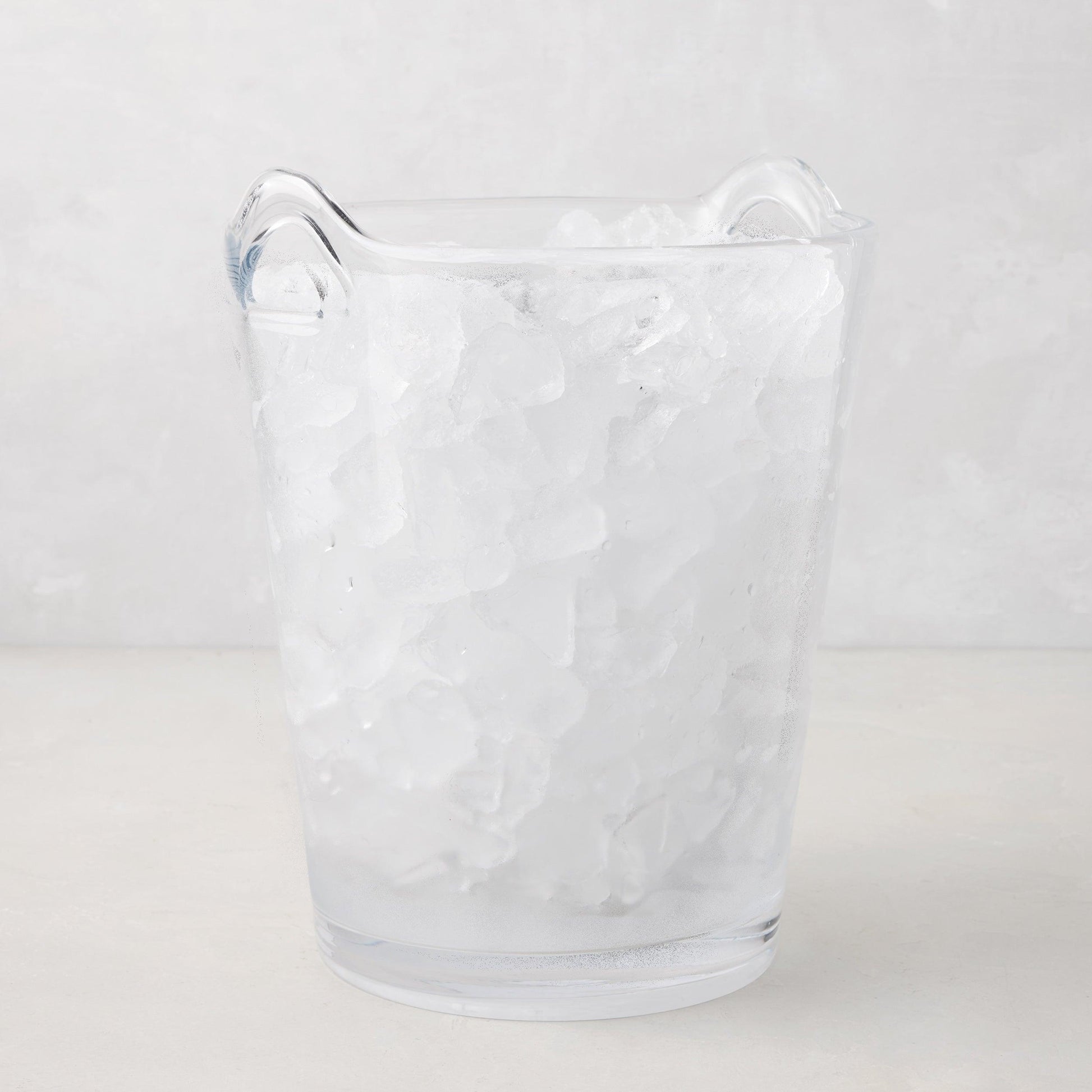 Large clear handblown glass ice bucket with two handles, filled with ice.