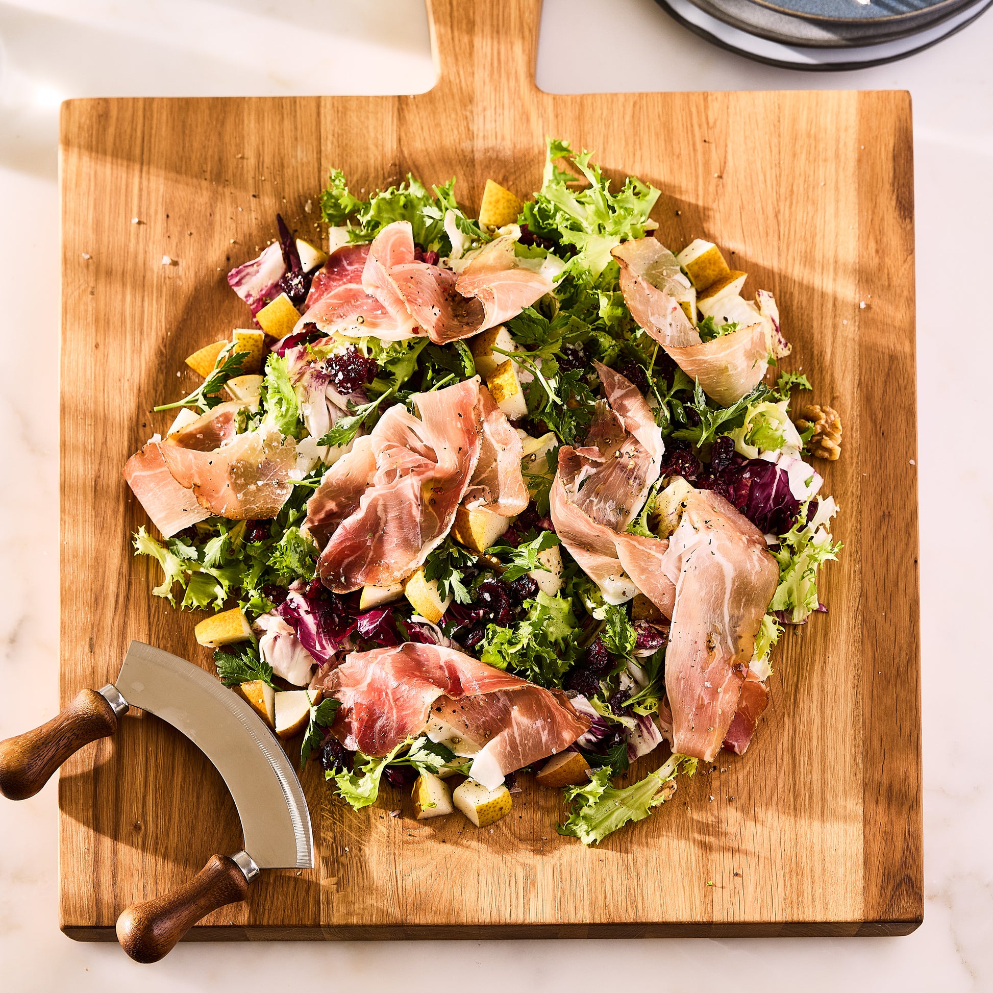 Large reclaimed oak wood cutting board bowl with salad, prosciutto, and mezzaluna blade.