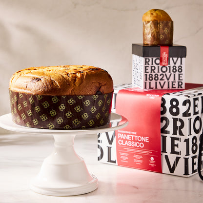 Large Olivieri classic panettone on a white cake stand next to branded black and white packaging boxes.