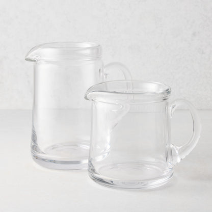 Two clear glass Oxford pitchers, one large and one small, with hand-molded rims and handles.