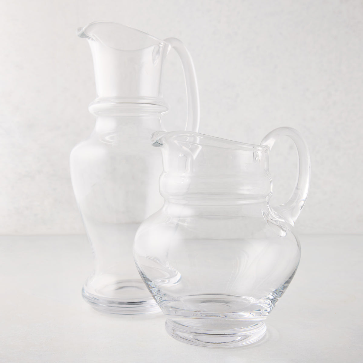 Two clear, hand-blown Sutton glass pitchers, one tall and one shorter, with unique shapes and handles.