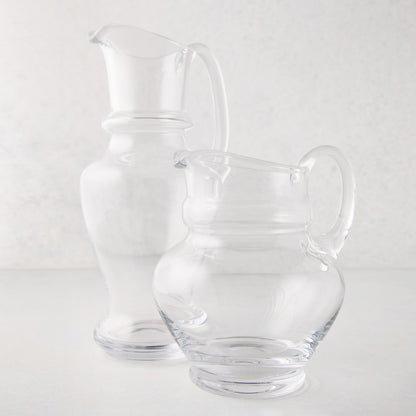 Two clear, hand-blown Sutton glass pitchers, one tall and one shorter, with unique shapes and handles.