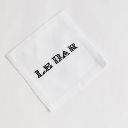 White linen cocktail napkin coaster with "LE BAR" hand-embroidered in black script.