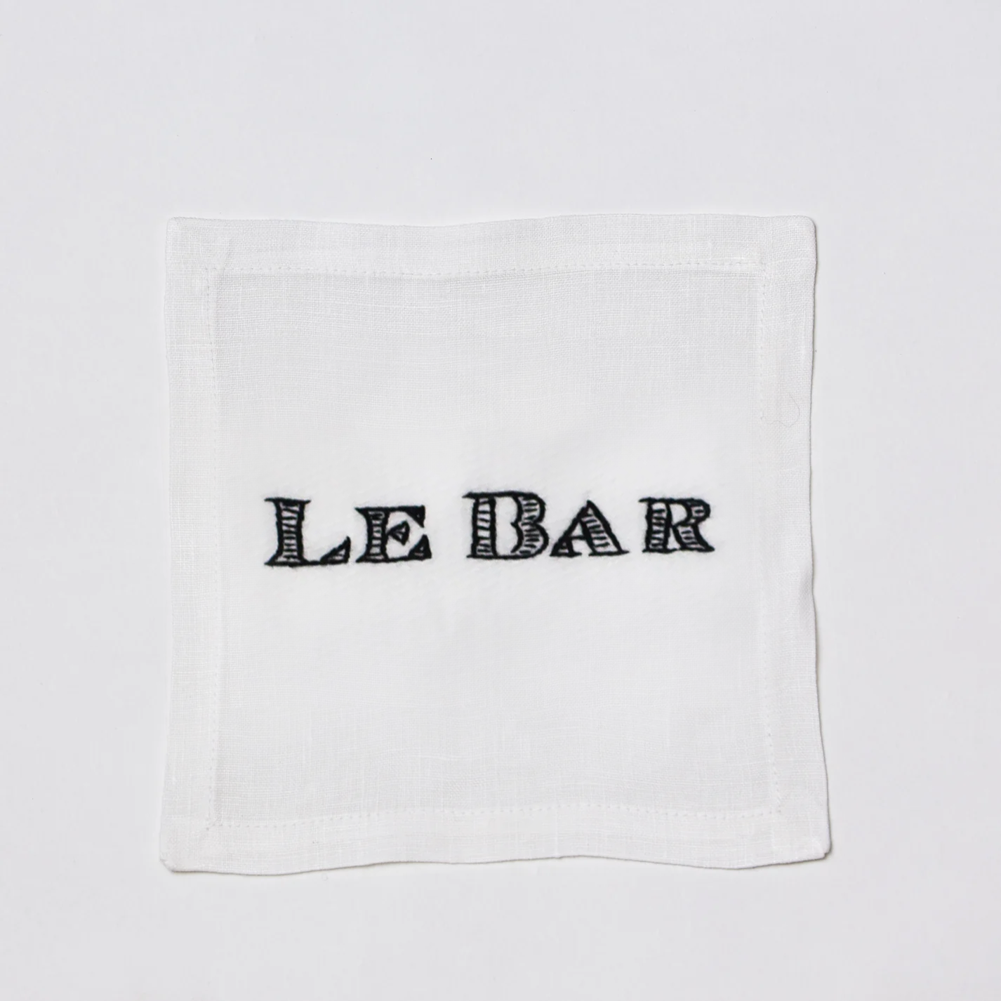White linen cocktail napkin coaster with "LE BAR" hand-embroidered in black.