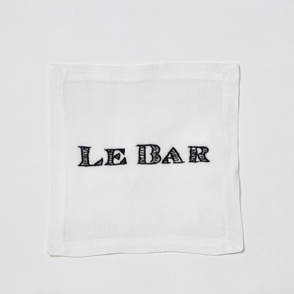 White linen cocktail napkin coaster with "LE BAR" hand-embroidered in black.