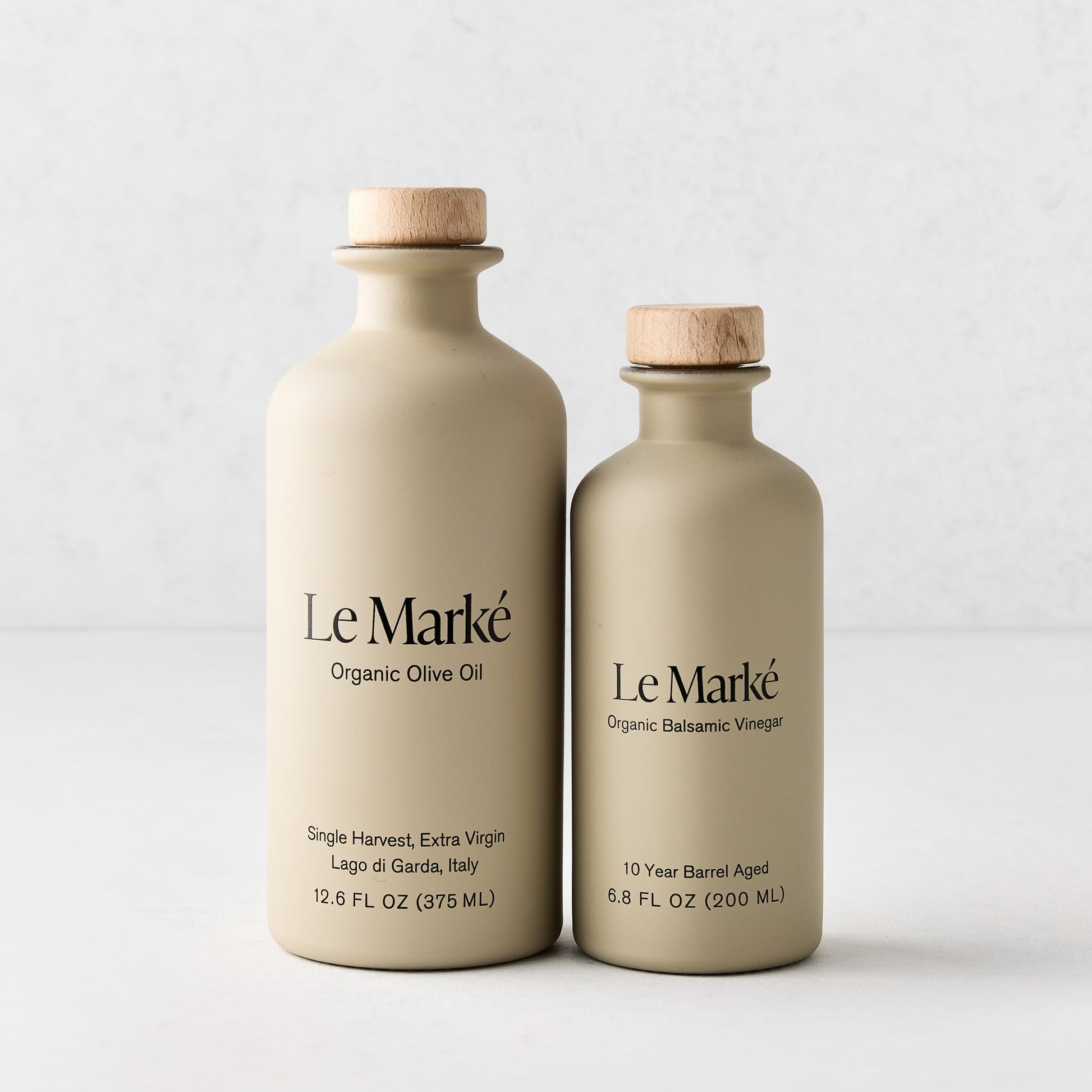 Le Marké balsamic vinegar and olive oil in matte beige glass bottles with wooden stoppers on a white background.
