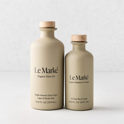 Two matte beige ceramic bottles of Le Marké olive oil and balsamic vinegar with wooden stoppers on a white surface.