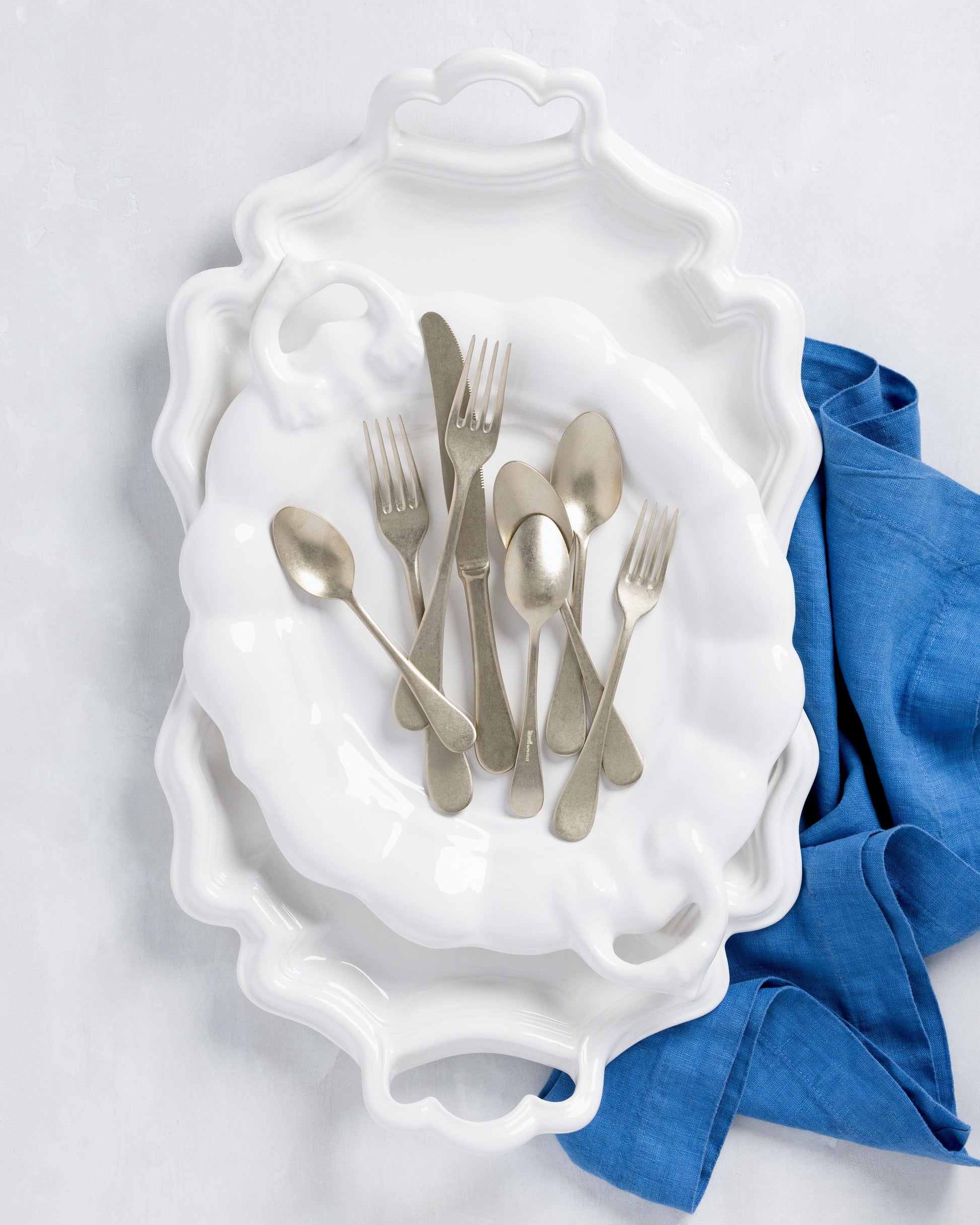 White ceramic oval serving platter with handles, stacked with a smaller platter, silverware, and blue napkin.