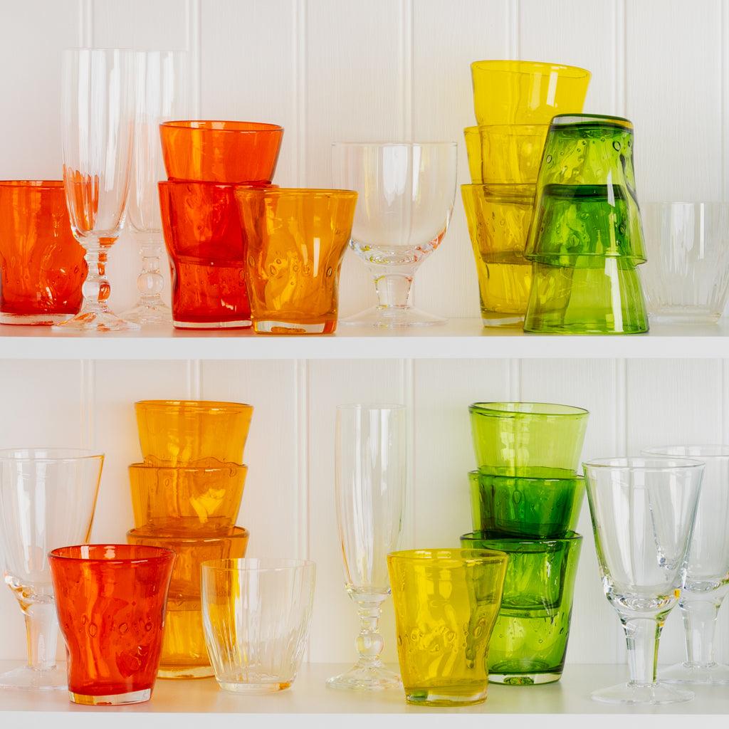 Light orange, yellow, green, and clear Henri mouth-blown glassware on white shelves.