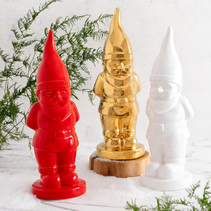 Three ceramic holiday gnomes: red, gold, and white, standing in faux snow with greenery.