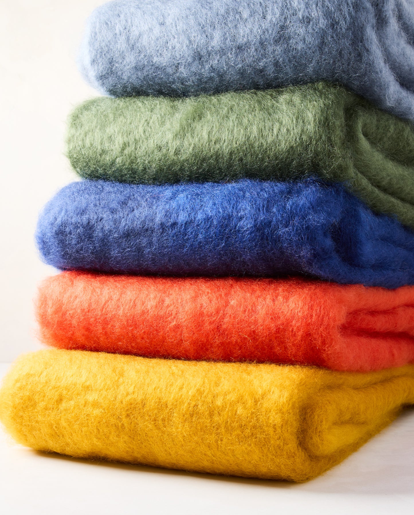 Stack of five folded mohair throws: blue, green, royal blue, orange, and yellow mustard.