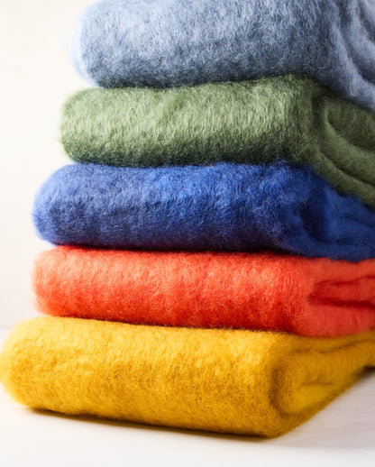 Stack of five folded mohair throws: blue, green, royal blue, orange, and yellow mustard.