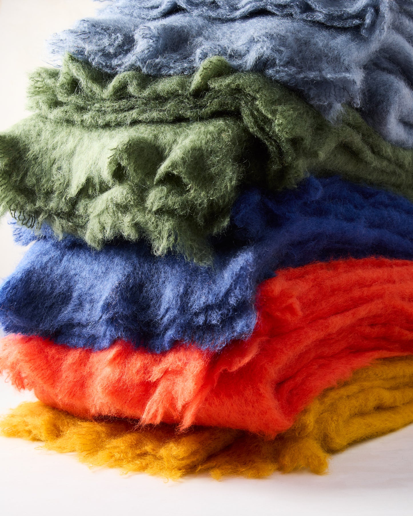 Stack of vibrant yellow, orange, blue, green, and grey mohair throws with a soft, fuzzy texture.