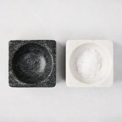 Black and white square marble pinch bowls, one filled with white salt, on a light background.