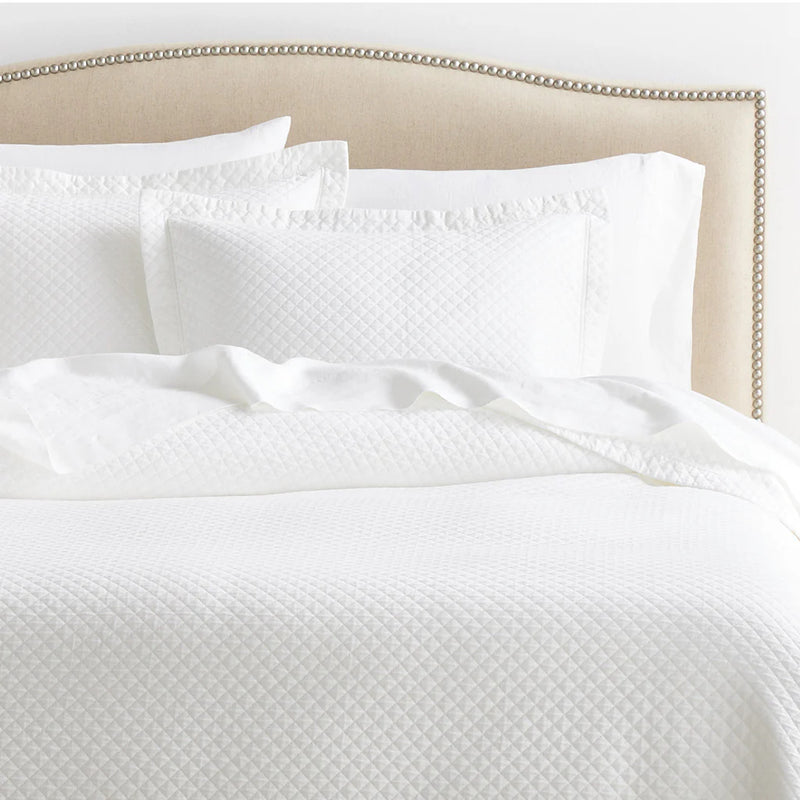 White matelassé quilt and matching pillow shams on a bed with a beige nailhead-trimmed upholstered headboard.