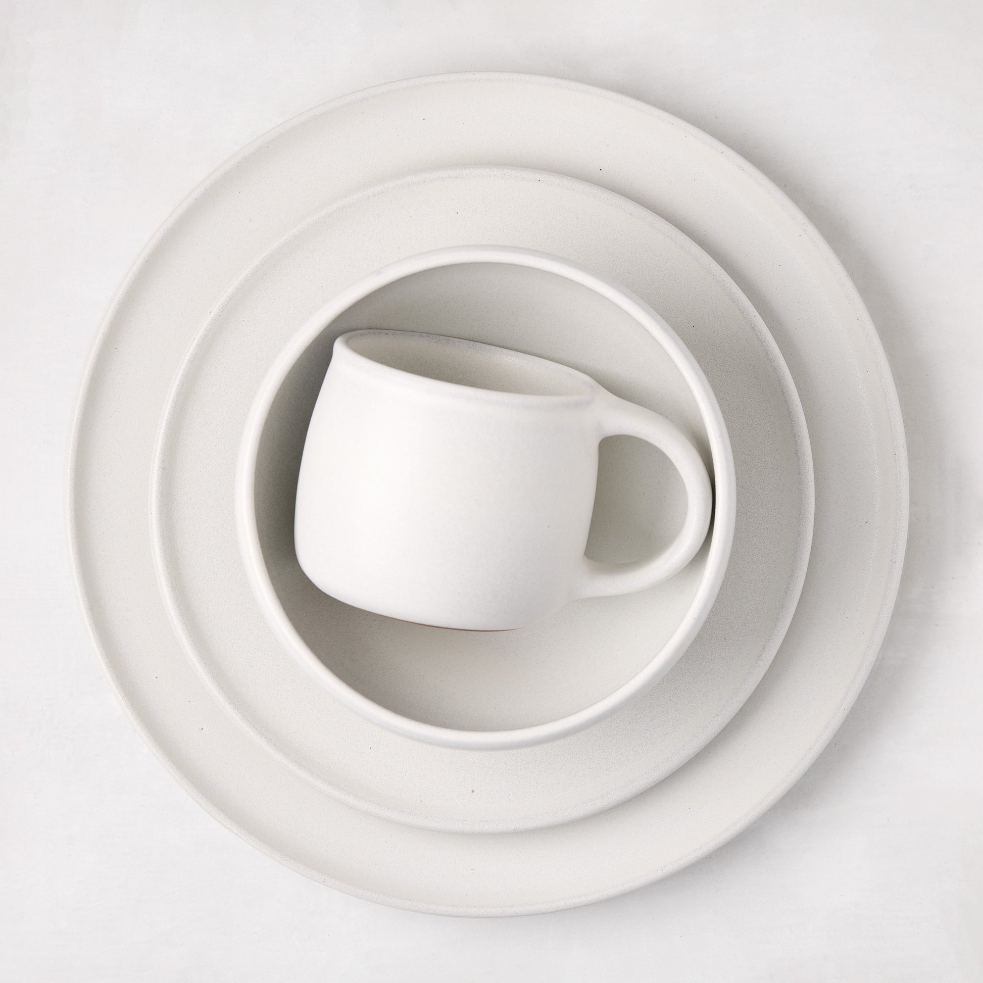 Matte white stoneware mug nested in a matching bowl, on two larger plates.