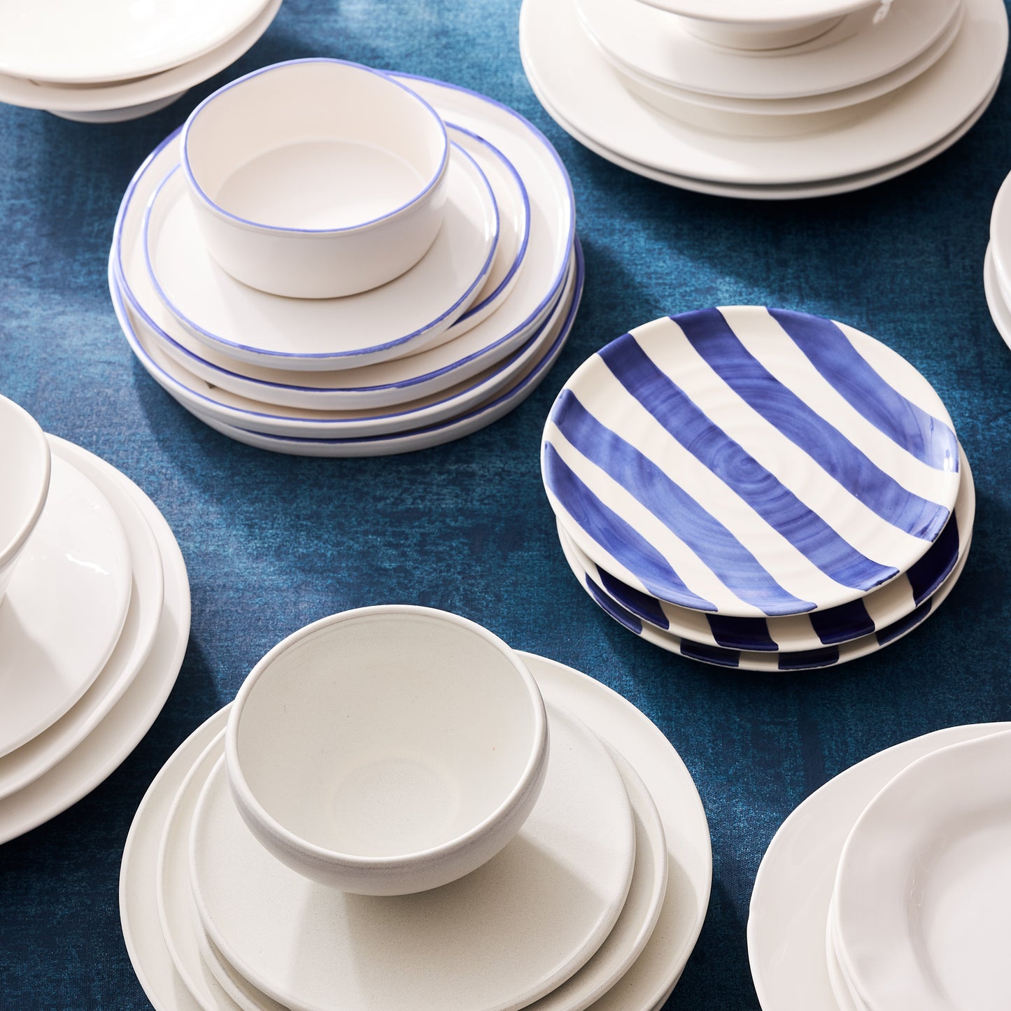 Matte white stoneware bowls and plates, some with blue rims or stripes, on a blue surface.