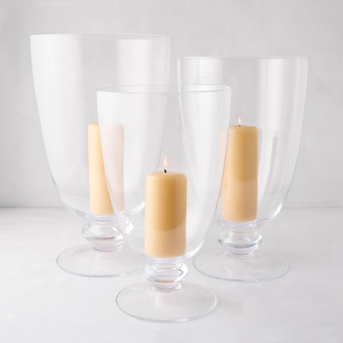 Three clear glass Mayfair hurricanes of varying sizes with decorative bases, holding lit cream candles.