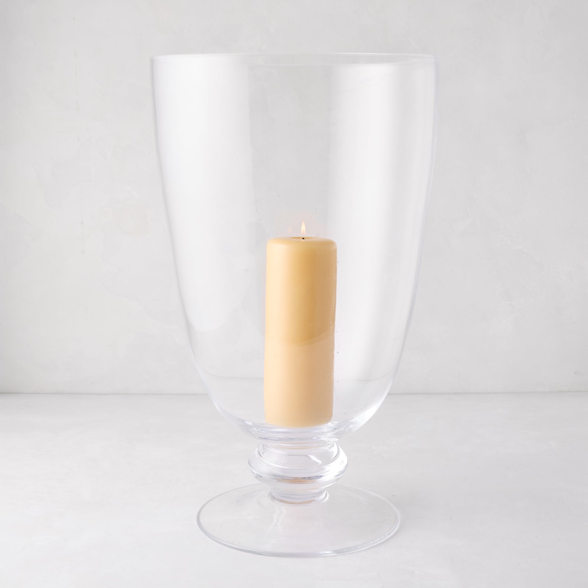 Large Mayfair clear glass hurricane with decorative base, holding a lit cream pillar candle.