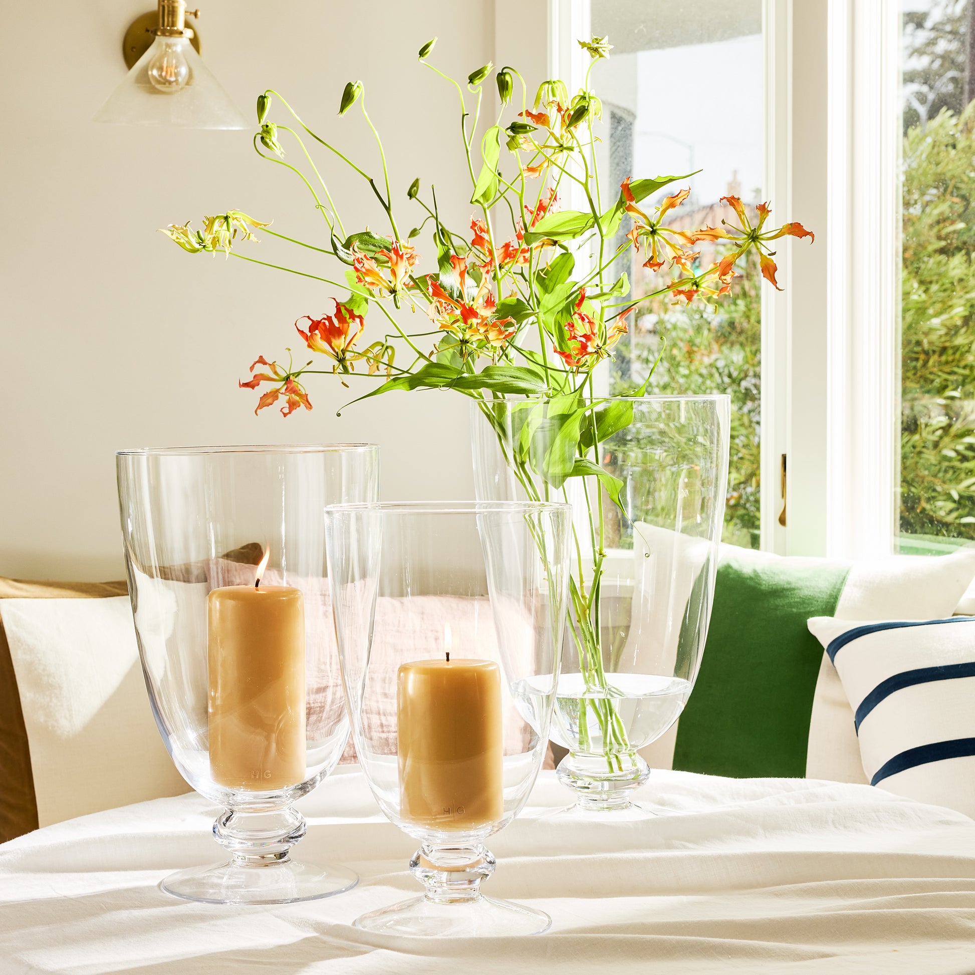 Medium Mayfair Glass Hurricane with two lit candles and a vase of orange flowers.